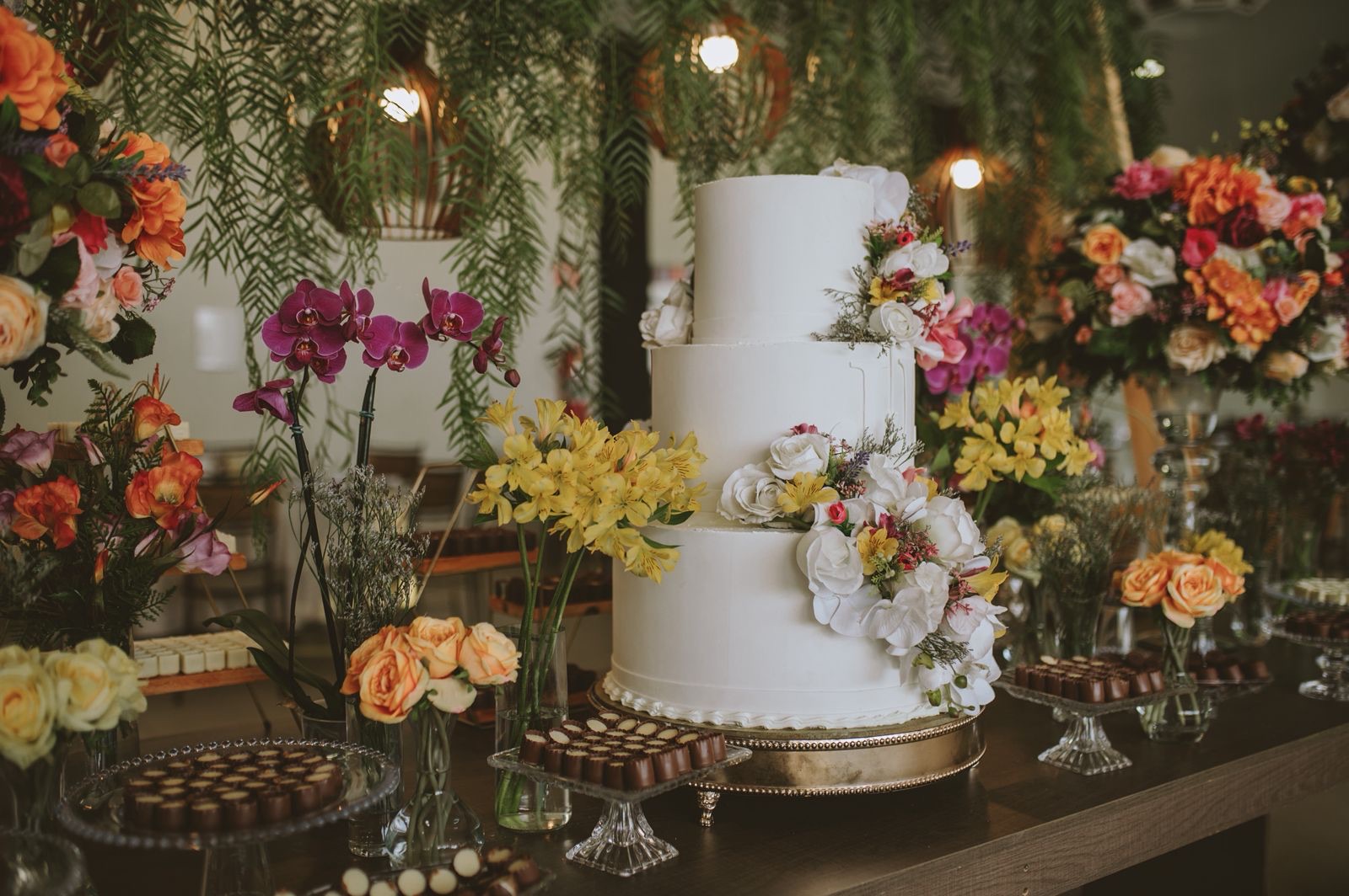Bolo de casamento de três andares cercado de flores e mesa de doces finos