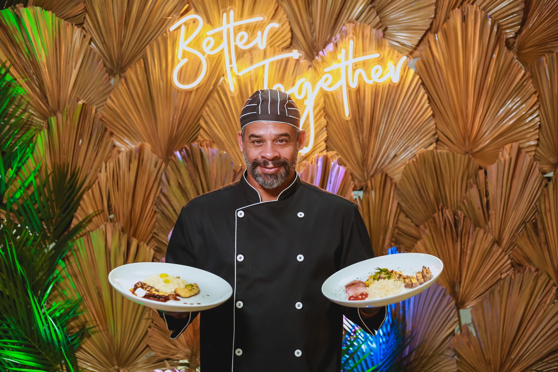 Chef Edson Brandão sorrindo com dois pratos, fundo de palmeiras secas e neon Better Together